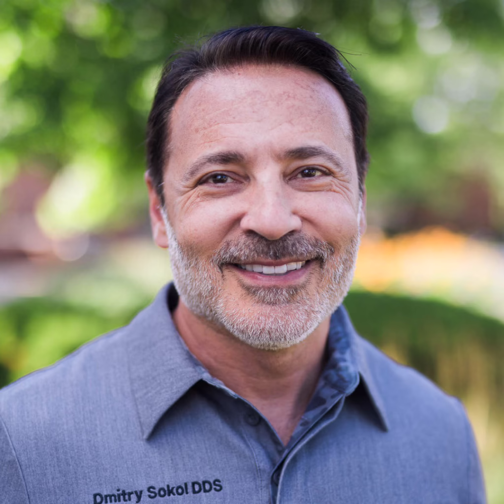 Smiling middle-aged man with short dark hair and gray beard, wearing a blue collared shirt outdoors.