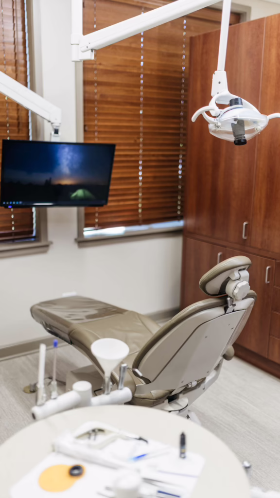 Dental examination chair in a clinic with overhead light, monitor, and wooden cabinets in the background.
