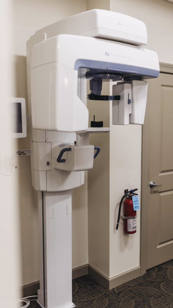 White panoramic dental X-ray machine on a stand in a clinic, with a wall-mounted monitor and a fire extinguisher nearby.