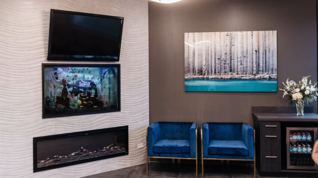 Lobby seating with two royal blue velvet chairs and a dark wood cabinet, a fresh flower arrangement, and wall art; left wall has a textured white pattern, right wall a large black-and-white photo above a teal-accent mural, and a fish tank mounted below a TV.