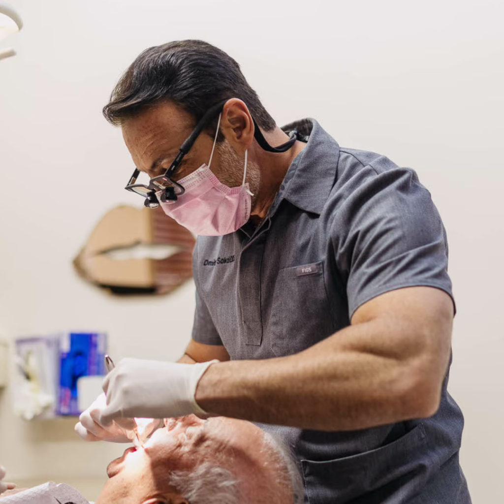 Dentist wearing pink mask and magnifying loupes examines a patient’s mouth while using dental tools; patient reclined in dental chair.