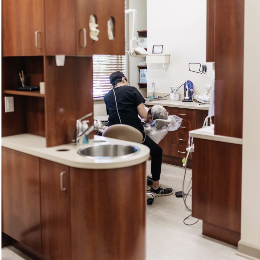 Dental hygienist in black scrubs attending to a patient reclined in a dental chair in a wood-paneled treatment room with cabinets and sink nearby.